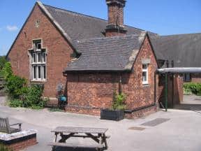 Red-brick building with pitched roof, chimney, picnic bench, and small garden on a sunny day.
