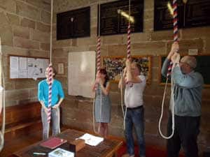 Four people ring striped church bell ropes inside a stone-walled room with notices on the walls.