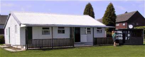Small white cricket pavilion with a scoreboard showing match totals on a grassy pitch.