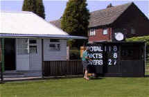 Person updating a cricket scorecard outside a small pavilion on a sunny day.