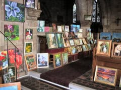 Many colourful paintings displayed inside a stone building with arched windows and a tiled floor.