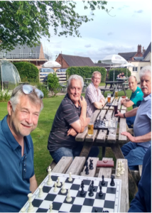 Six men sit at picnic tables outdoors, playing chess and enjoying drinks on a sunny day.