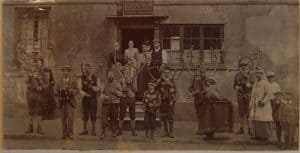 A sepia photograph of people with antlers standing in front of a building, with others observing from the doorway.